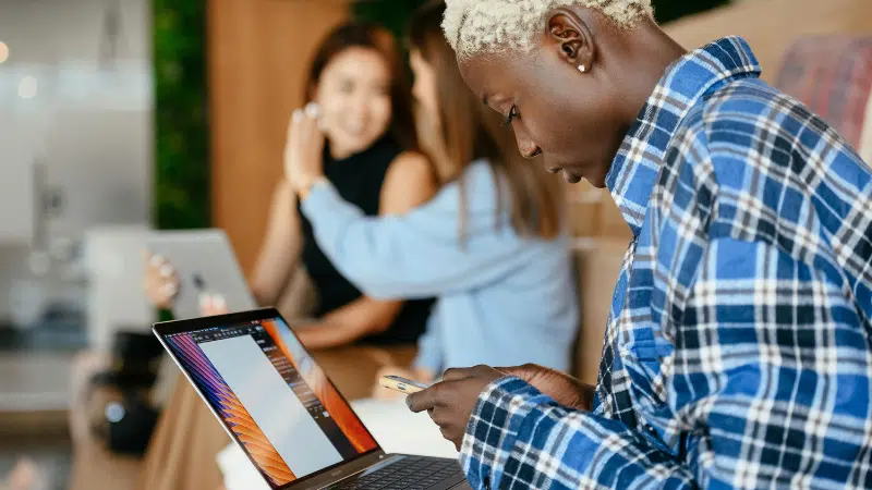 A person in a plaid shirt uses a smartphone and laptop, possibly reviewing Amazon Music Insights, while two people with laptops talk in the background.