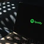 A laptop displaying the Spotify logo on its screen sits on a dark surface, surrounded by dappled light patterns that add an atmospheric touch.