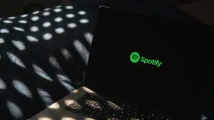 A laptop displaying the Spotify logo on its screen sits on a dark surface, surrounded by dappled light patterns that add an atmospheric touch.