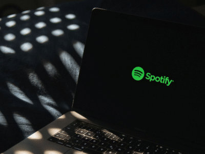 A laptop displaying the Spotify logo on its screen sits on a dark surface, surrounded by dappled light patterns that add an atmospheric touch.