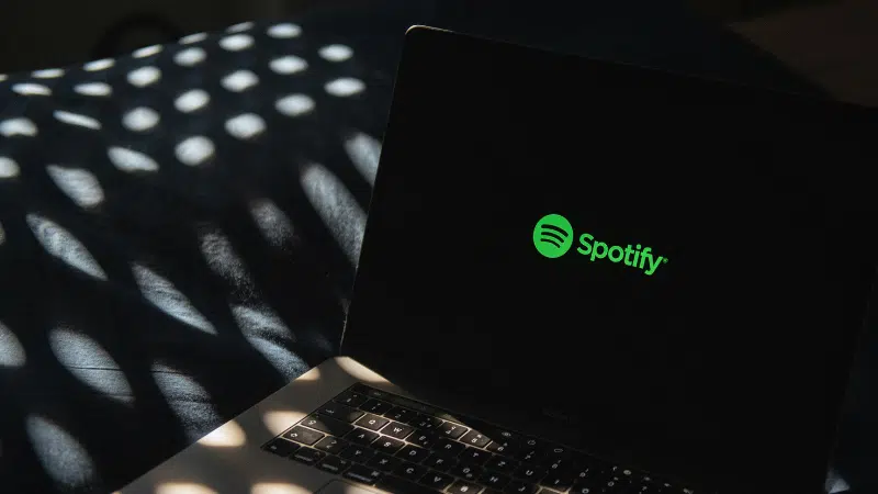 A laptop displaying the Spotify logo on its screen sits on a dark surface, surrounded by dappled light patterns that add an atmospheric touch.