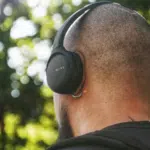 A person with a shaved head and backpack checks their email while wearing black Sony over-ear headphones outdoors, with trees in the background.