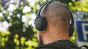 A person with a shaved head and backpack checks their email while wearing black Sony over-ear headphones outdoors, with trees in the background.