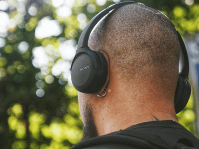 A person with a shaved head and backpack checks their email while wearing black Sony over-ear headphones outdoors, with trees in the background.