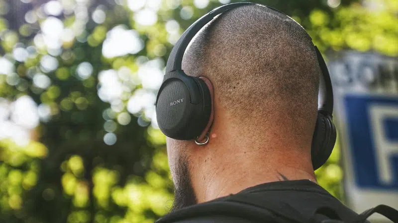 A person with a shaved head and backpack checks their email while wearing black Sony over-ear headphones outdoors, with trees in the background.