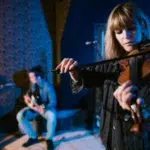A woman plays the violin while two other musicians play guitars in the background, all performing in a dimly lit indoor space—showcasing how to build a scalable music business through captivating live performances.