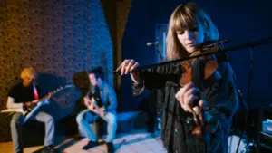 A woman plays the violin while two other musicians play guitars in the background, all performing in a dimly lit indoor space—showcasing how to build a scalable music business through captivating live performances.