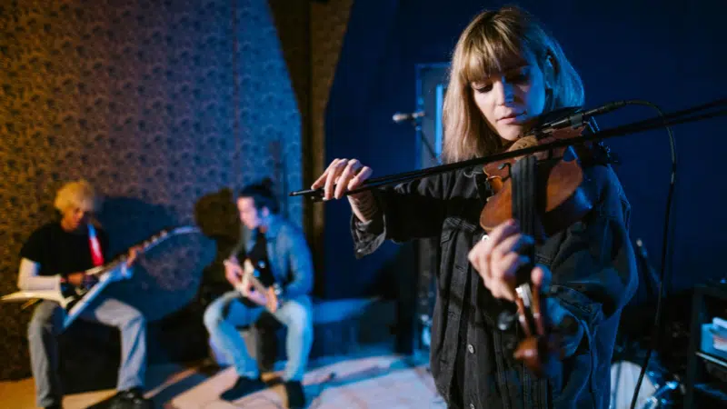 A woman plays the violin while two other musicians play guitars in the background, all performing in a dimly lit indoor space—showcasing how to build a scalable music business through captivating live performances.