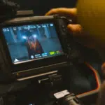 A person operates a video camera in perfect sync, capturing footage of someone standing in a hallway as displayed on the camera’s monitor screen.