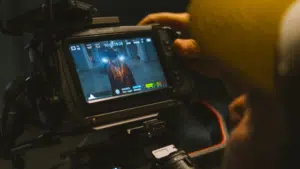 A person operates a video camera in perfect sync, capturing footage of someone standing in a hallway as displayed on the camera’s monitor screen.