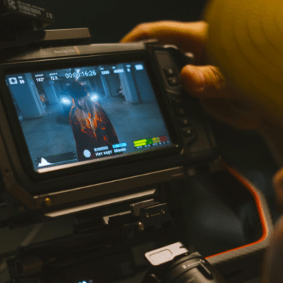 A person operates a video camera in perfect sync, capturing footage of someone standing in a hallway as displayed on the camera’s monitor screen.