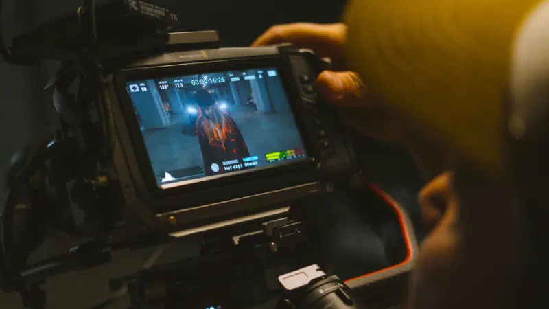 A person operates a video camera in perfect sync, capturing footage of someone standing in a hallway as displayed on the camera’s monitor screen.