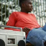 Two people sit outdoors by a metal fence, with a silver boombox placed in the foreground.
