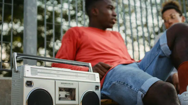 Two people sit outdoors by a metal fence, with a silver boombox placed in the foreground.