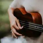 A person celebrating Hispanic heritage wears a beige shirt and strums a wooden ukulele outdoors, with greenery blurred in the background.