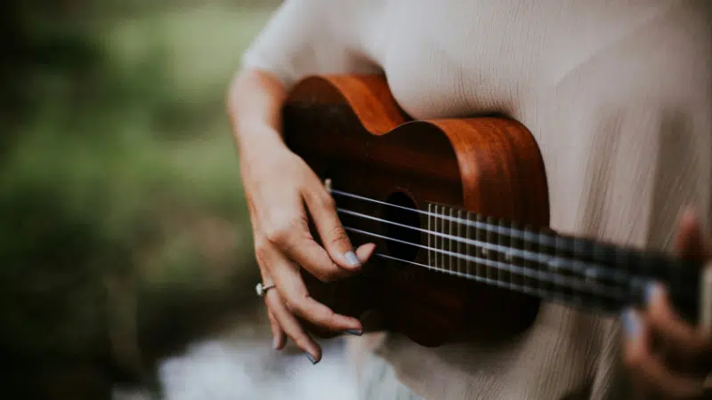 A person celebrating Hispanic heritage wears a beige shirt and strums a wooden ukulele outdoors, with greenery blurred in the background.