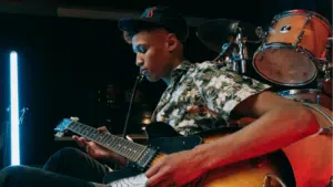 A young man wearing a floral shirt and cap plays an electric guitar on stage, with drums and stage lights in the background, celebrating the release of new music.