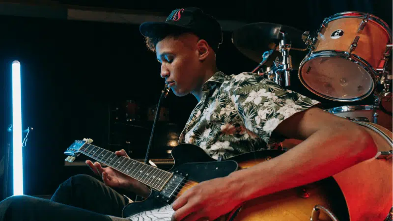 A young man wearing a floral shirt and cap plays an electric guitar on stage, with drums and stage lights in the background, celebrating the release of new music.