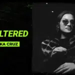 Black and white image with text "Symphonic Unfiltered with Chesk Cruz" next to a woman wearing sunglasses and a patterned coat, standing in a dimly lit setting.