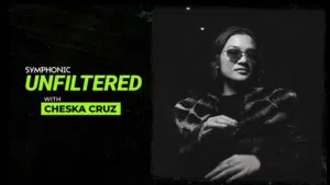 Black and white image with text "Symphonic Unfiltered with Chesk Cruz" next to a woman wearing sunglasses and a patterned coat, standing in a dimly lit setting.