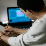 A person wearing a beanie writes on sheet music with a pencil at a desk, while SongTools audio software is open on their laptop in the background.