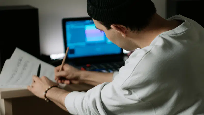 A person wearing a beanie writes on sheet music with a pencil at a desk, while SongTools audio software is open on their laptop in the background.
