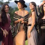 Five women stand outdoors in a grassy area, wearing dresses and patterned shawls, as if pausing mid-playlist for a photo, with trees and mountains visible in the background.