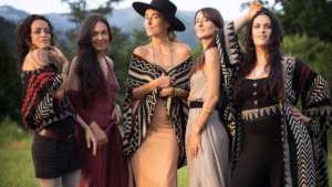 Five women stand outdoors in a grassy area, wearing dresses and patterned shawls, as if pausing mid-playlist for a photo, with trees and mountains visible in the background.