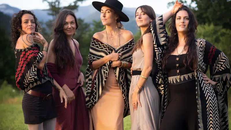 Five women stand outdoors in a grassy area, wearing dresses and patterned shawls, as if pausing mid-playlist for a photo, with trees and mountains visible in the background.