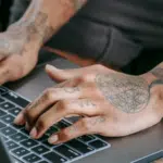 Tattooed hands typing on a laptop keyboard.