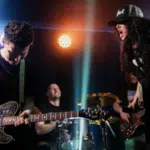 A rock band performs on stage with a guitarist, drummer, bassist, and a vocalist in a hat singing passionately under colorful stage lights, drawing the crowd into their energetic fan funnel.