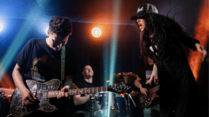 The Independent Artist Funnel: How to Think Like a Brand A rock band performs on stage with a guitarist, drummer, bassist, and a vocalist in a hat singing passionately under colorful stage lights, drawing the crowd into their energetic fan funnel.