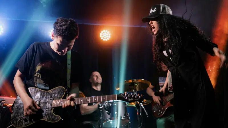 A rock band performs on stage with a guitarist, drummer, bassist, and a vocalist in a hat singing passionately under colorful stage lights, drawing the crowd into their energetic fan funnel.