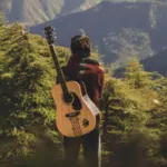 Person standing in a forested area with a guitar slung over their back, facing away and gazing at distant mountains under daylight—a peaceful scene that echoes the quiet individuality found in micro-markets.