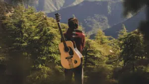 Person standing in a forested area with a guitar slung over their back, facing away and gazing at distant mountains under daylight—a peaceful scene that echoes the quiet individuality found in micro-markets.