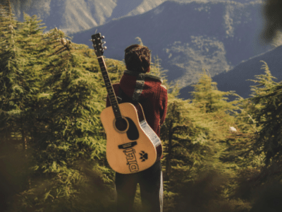 Person standing in a forested area with a guitar slung over their back, facing away and gazing at distant mountains under daylight—a peaceful scene that echoes the quiet individuality found in micro-markets.