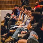 A group of people sit in rows of seats in an auditorium, attentively watching or listening to an event or presentation.