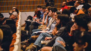 A group of people sit in rows of seats in an auditorium, attentively watching or listening to an event or presentation.