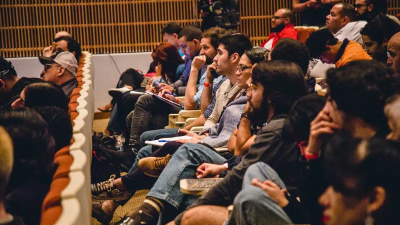 A group of people sit in rows of seats in an auditorium, attentively watching or listening to an event or presentation.