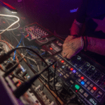 A distributor expertly operates a variety of electronic music equipment and synthesizers, illuminated by colorful stage lighting.
