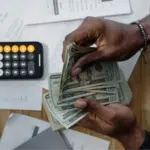 A person counts US dollar bills at a desk with a calculator, scattered financial documents, and the SplitShare app open on their screen.