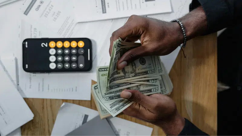 A person counts US dollar bills at a desk with a calculator, scattered financial documents, and the SplitShare app open on their screen.