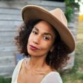A woman with curly hair wearing a wide-brimmed tan hat and a white top stands outdoors in front of a wooden wall, looking at the camera.