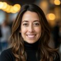 A woman with long brown hair, wearing a black turtleneck, smiles at the camera indoors with warm, blurred lights in the background.