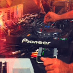 A person operates a Pioneer DJ controller with various cables connected, next to a laptop running Spotify, in a dimly lit environment with colorful lights.