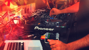 Big News: Spotify Now Integrates with DJ Software! A person operates a Pioneer DJ controller with various cables connected, next to a laptop running Spotify, in a dimly lit environment with colorful lights.