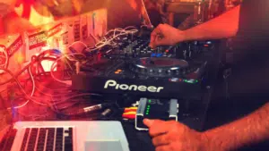 A person operates a Pioneer DJ controller with various cables connected, next to a laptop running Spotify, in a dimly lit environment with colorful lights.