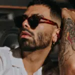 A man wearing sunglasses and a white shirt sits in front of speakers, showing a tattoo on his arm and touching his head, channeling the bold style often seen among influential Hispanic artists.
