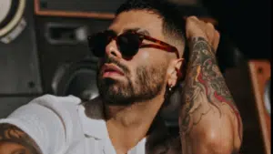 A man wearing sunglasses and a white shirt sits in front of speakers, showing a tattoo on his arm and touching his head, channeling the bold style often seen among influential Hispanic artists.