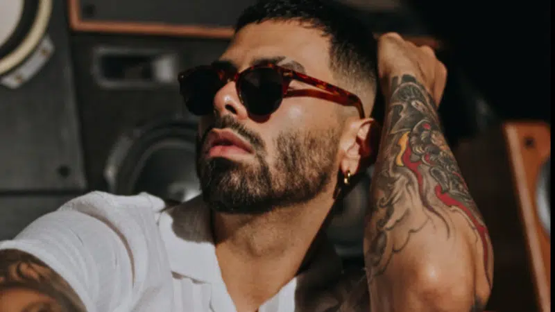 A man wearing sunglasses and a white shirt sits in front of speakers, showing a tattoo on his arm and touching his head, channeling the bold style often seen among influential Hispanic artists.
